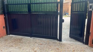 Een gesloten zwart metalen hek met gaas bovenop, leidend naar een verhard gebied met verspreide bladeren.