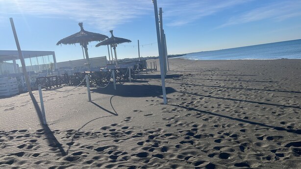Een strand met parasols, tafels en een gebouw met een glazen gevel.