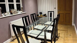 Een eetruimte met een glazen tafel, omringd door stoelen, een radiator, een raam met decoratieve objecten en een deur.
