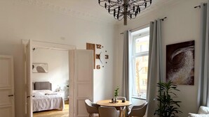 A spacious room with a wooden floor, a round dining table with chairs, a bed with a grey headboard, a white sofa, and a large window with curtains. - Apartment Stella – Elegant Historic Apartment in the City Center (Kassel)