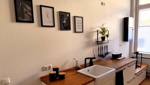 A modern kitchen with a wooden countertop, white cabinets, a sink, and a black faucet. There are framed pictures on the wall, a potted plant, and a window with blinds. - Apartment Stella – Elegant Historic Apartment in the City Center (Kassel)