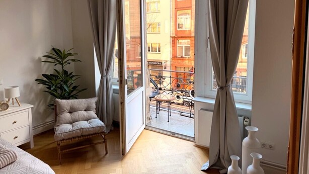 A bedroom with a large window, a bed, a nightstand, a chair, and a potted plant. - Apartment Stella – Elegant Historic Apartment in the City Center (Kassel)