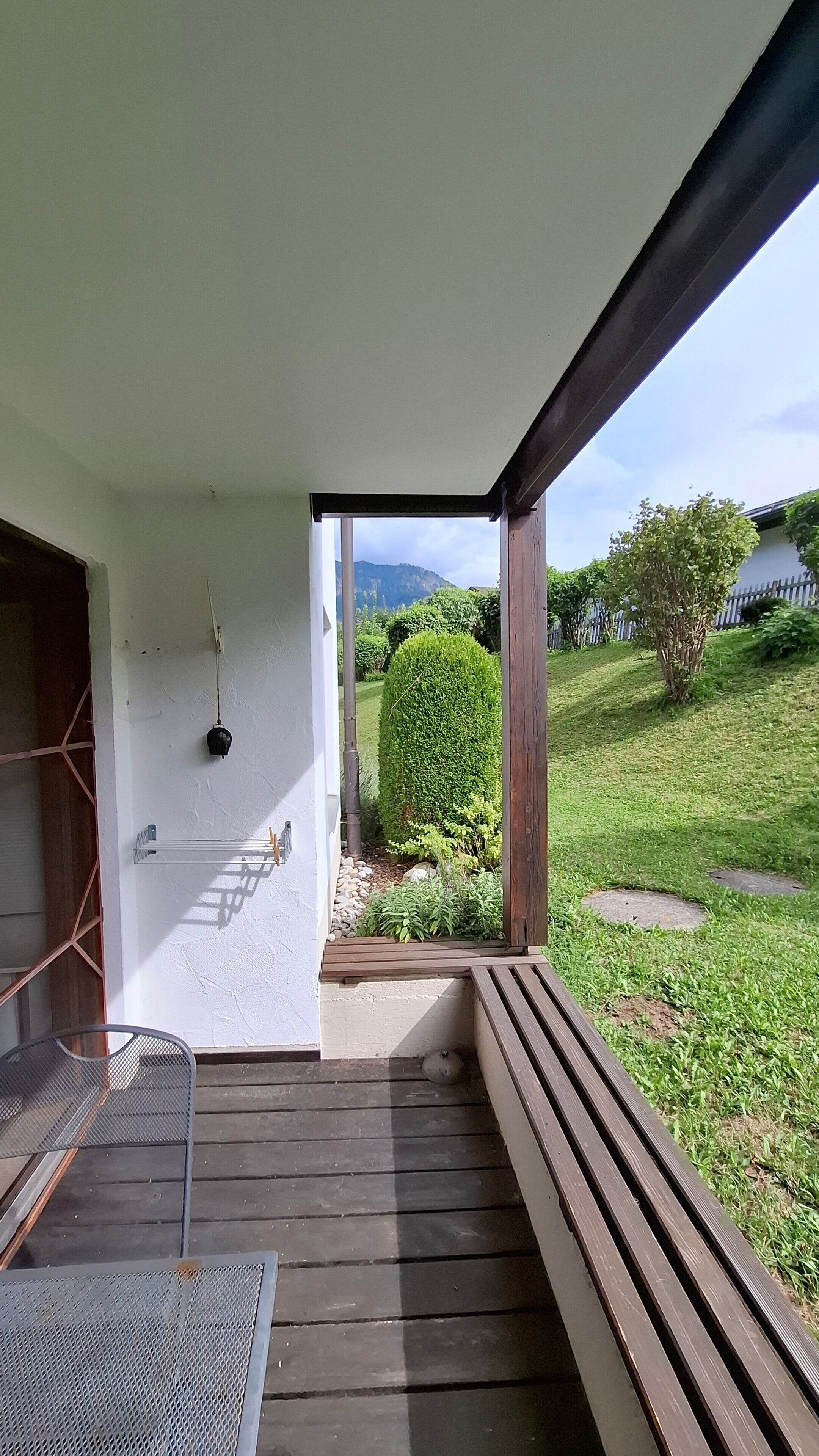 Eine Holzterrasse mit einer Bank, einer Hängeleuchte und Blick auf einen gepflegten Garten mit Hecke und Rasen.