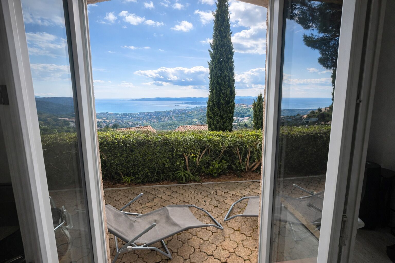 A view from a sliding glass door to an outdoor patio with a metal chair and a hedge.