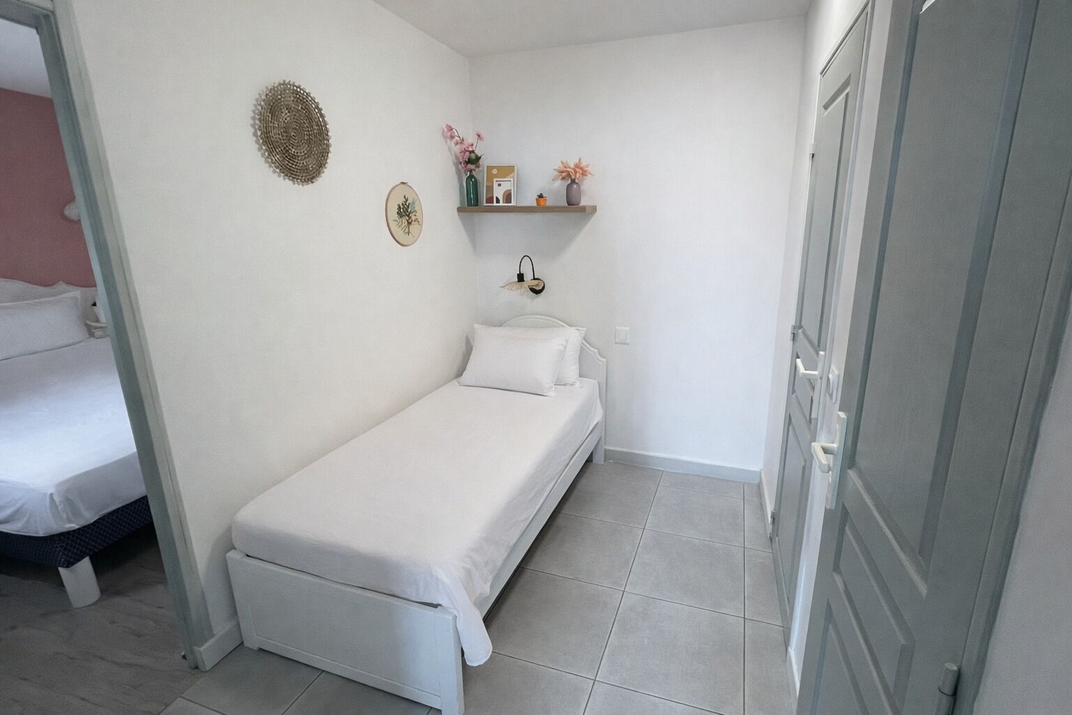 Een kleine, verzorgde slaapkamer met een eenpersoonsbed, een plankje met decoratieve spullen, en een deur naar een andere kamer.