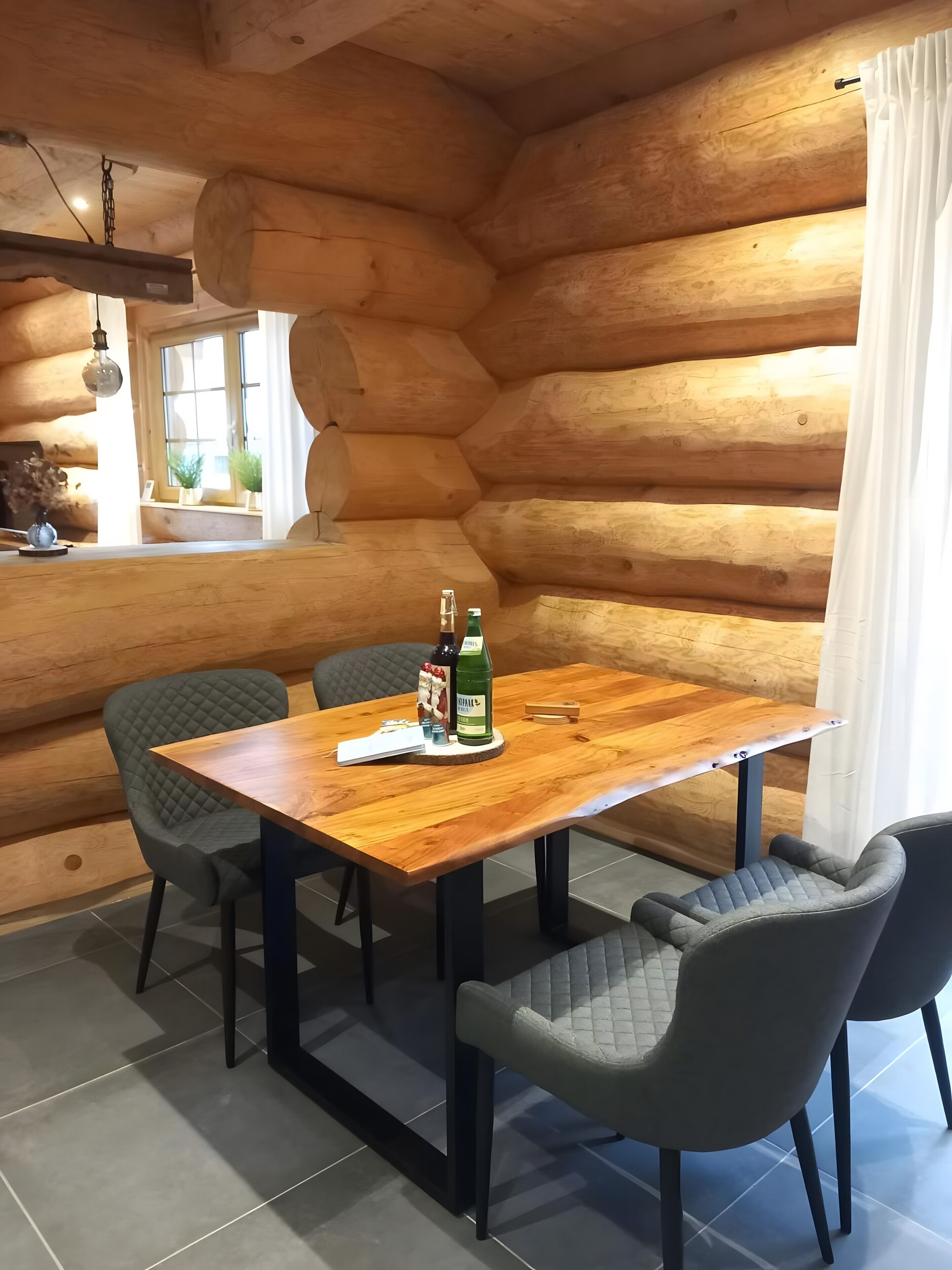 Interieur van een houten blokhut met een eettafel, vier stoelen, een hanglamp en een raam met planten.