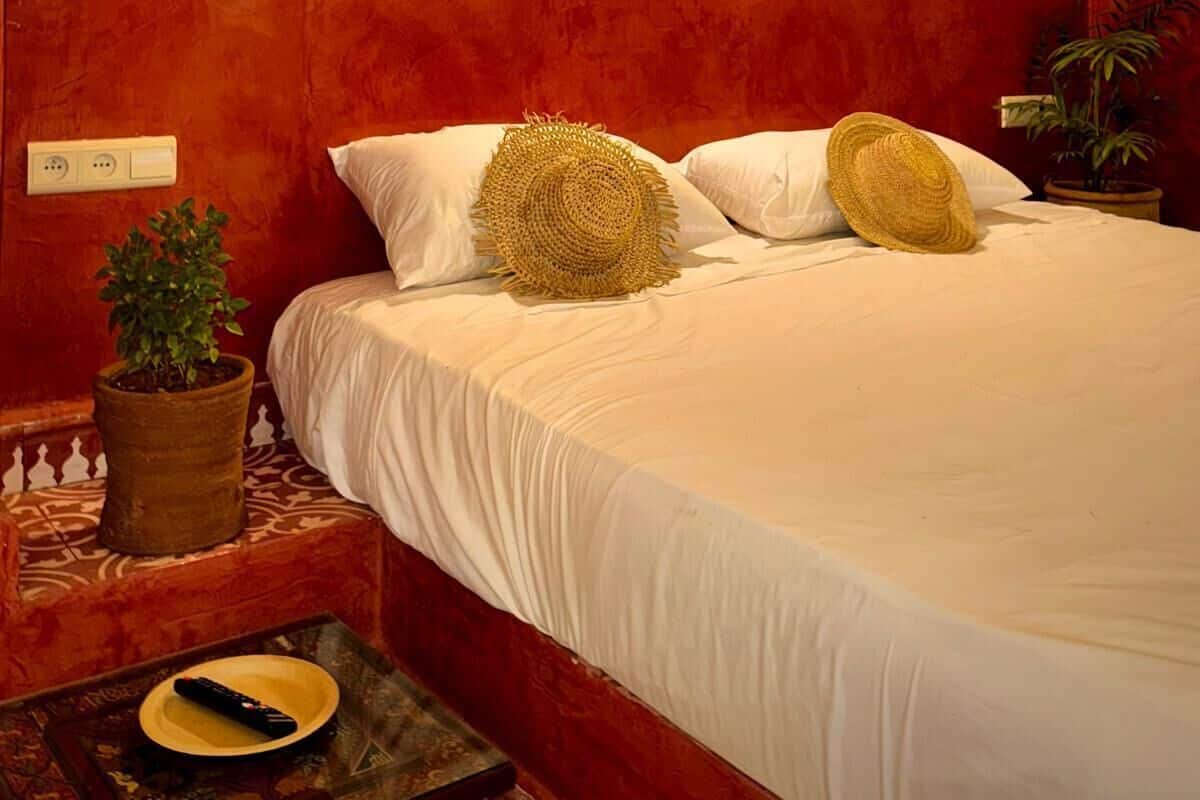 Une chambre à coucher avec une tête de lit dont le mur est peint en rouge, une lampe de chevet, une plante en pot et une télécommande posée sur la table de chevet.