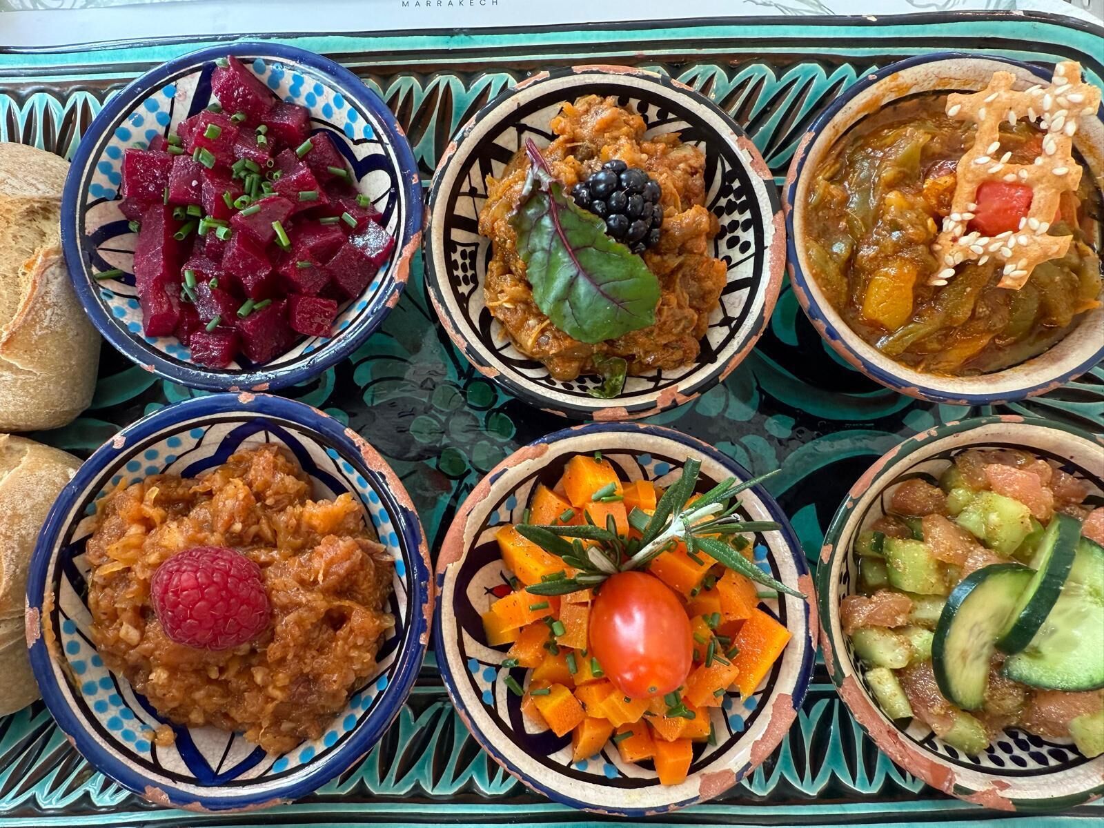 A tray with various dishes including beetroot salad, a berry compote, a vegetable stew, a carrot salad, a hummus with a raspberry, and a cucumber salad.