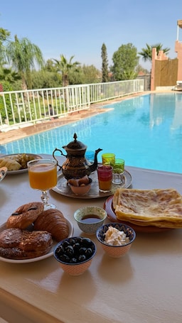 Una mesa de desayuno con pasteles, un calentador de té, vasos y vistas a una piscina.