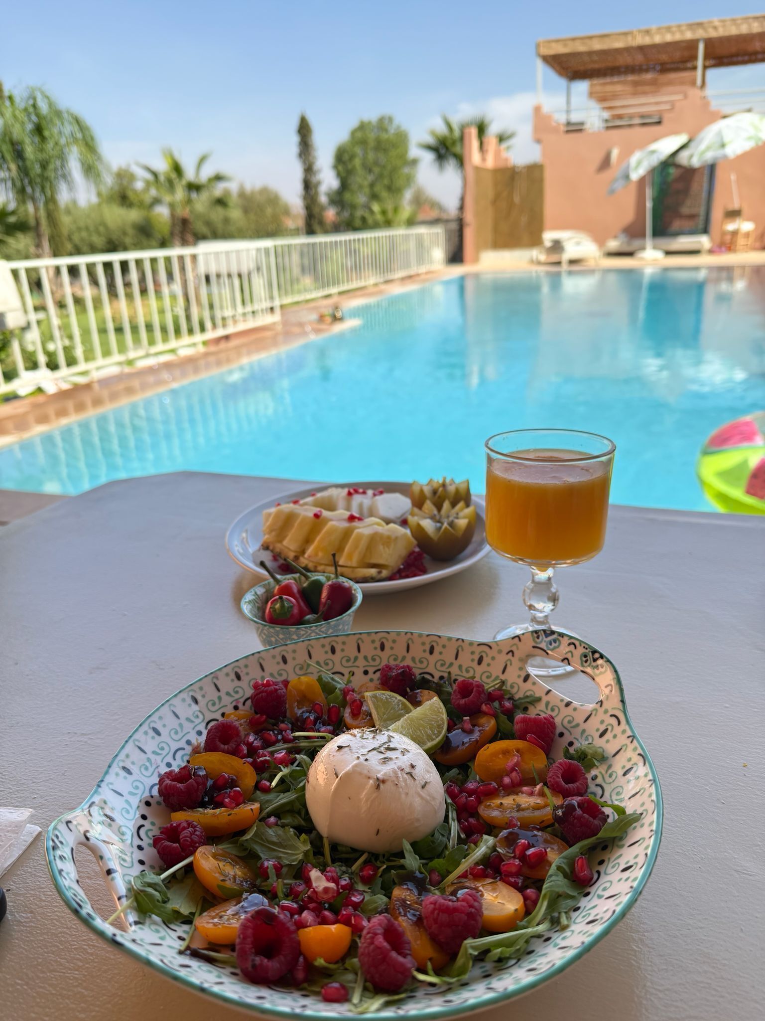 Una mesa de comedor con ensalada, un vaso de jugo y un plato de fruta al lado de una piscina.