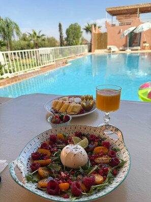 A dining table with a salad, a glass of juice, and a plate of fruit by a poolside. - Domaine JJ (Sidi Abdallah Ghiat)