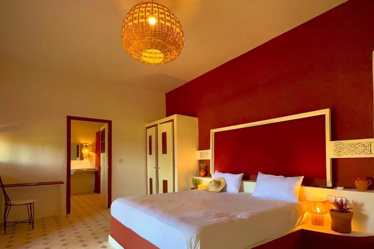 Une chambre avec un grand lit, un mur d’accent de couleur rouge, un sol à motifs géométriques et un luminaire suspendu.