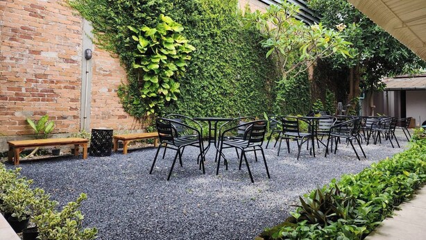 Un área de estar al aire libre con mesas y sillas, rodeada de piso de grava y un muro de ladrillo cubierto de vegetación.
