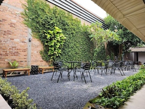 An outdoor seating area with tables and chairs, surrounded by a gravel floor and a brick wall covered in greenery. - RYB Hostel Phuket (Phuket)