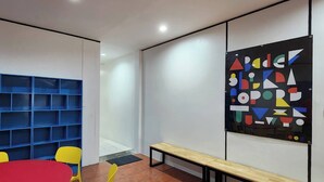 Una habitación con una mesa roja, sillas amarillas, un estante azul, un banco de madera y una obra de arte geométrica de colores en la pared.