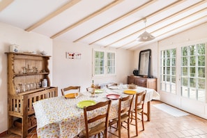 Un coin repas avec une table dressée pour quatre personnes, des chaises en bois, un buffet contenant de la verrerie et une grande fenêtre donnant sur un paysage verdoyant.