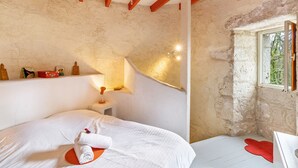 A bedroom with a bed, a small table, a window, and a wooden ceiling beam.