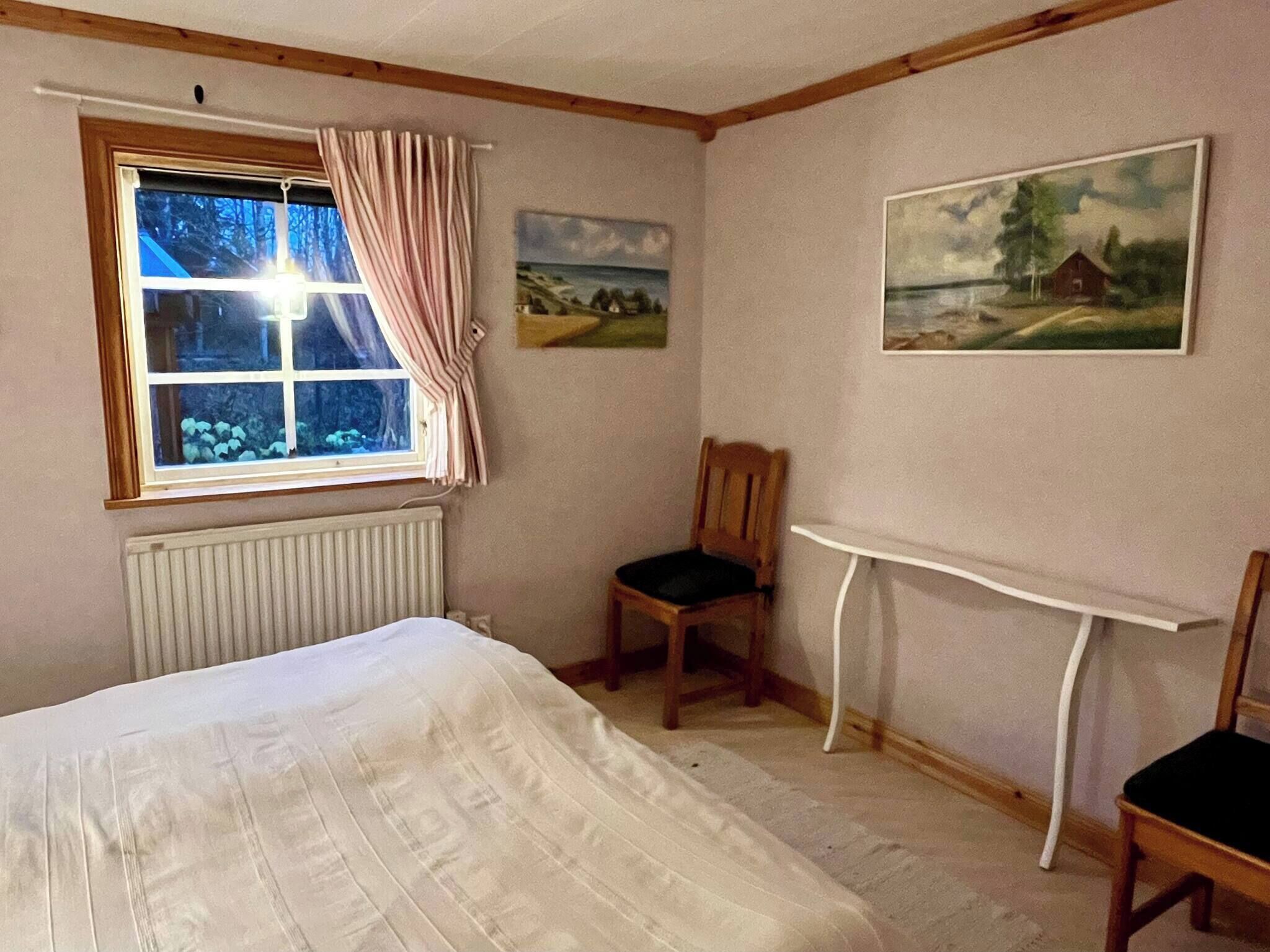 A room with a bed, a chair, a table, a window with curtains, and two paintings on the wall.
