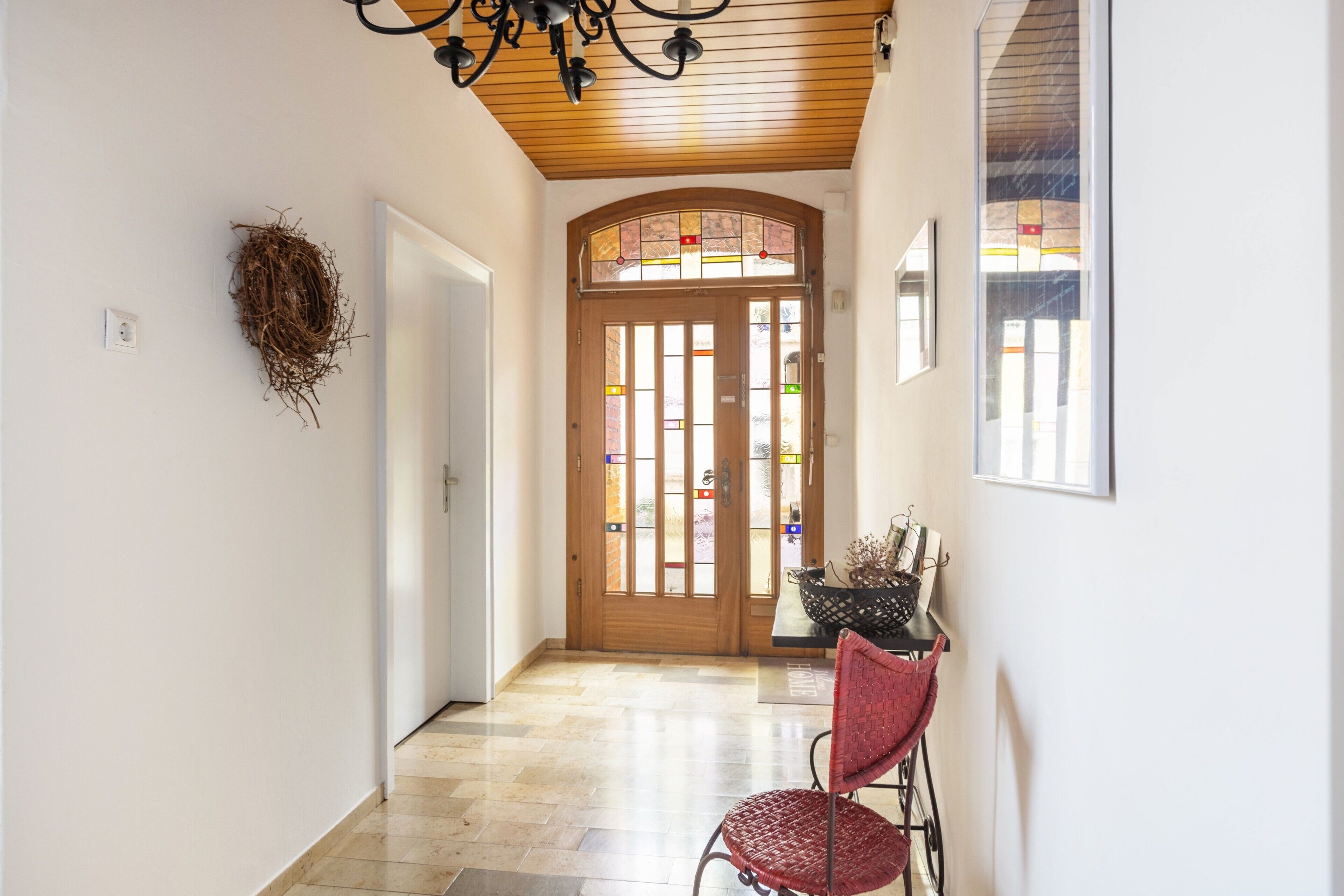 A hallway with a wooden ceiling, a door with stained glass, a wall-mounted nest, and a chair with a woven seat.