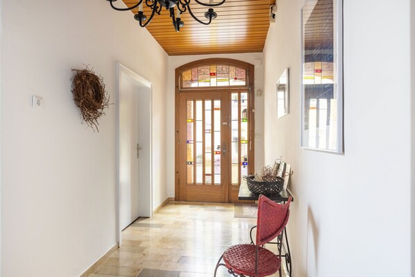 Un couloir avec un plafond en bois, une porte ornée de vitraux, un nid fixé au mur et une chaise avec un siège en osier tressé.