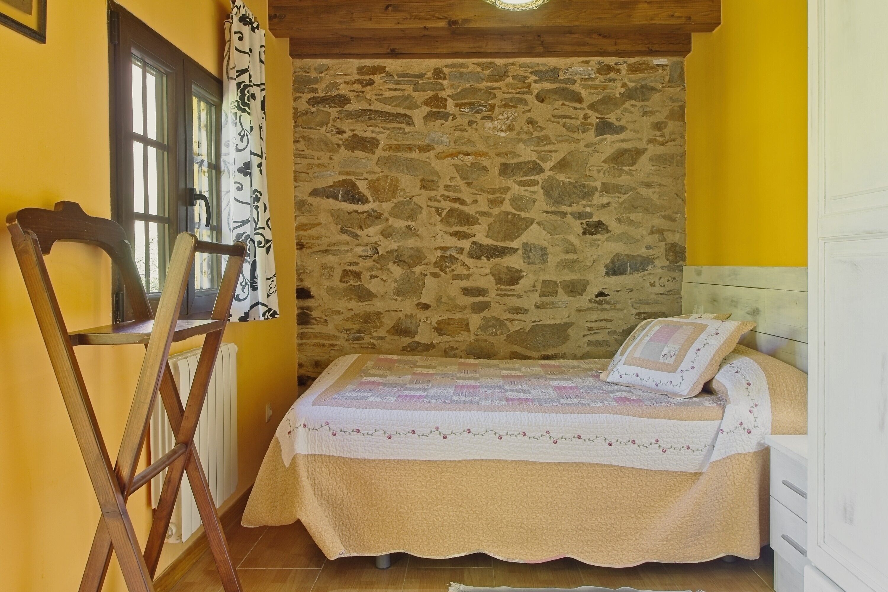 Une chambre avec un mur en pierre, un lit recouvert d’une courtepointe, une chaise et une fenêtre avec des rideaux.
