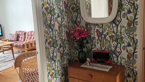 Una habitación con papel tapiz estampado, un aparador de madera con cajones, un espejo y una silla de mimbre.