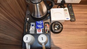 Uma cozinha compacta com chaleira de aço inoxidável, cafeteira, micro-ondas, controle remoto e um conjunto de canecas.