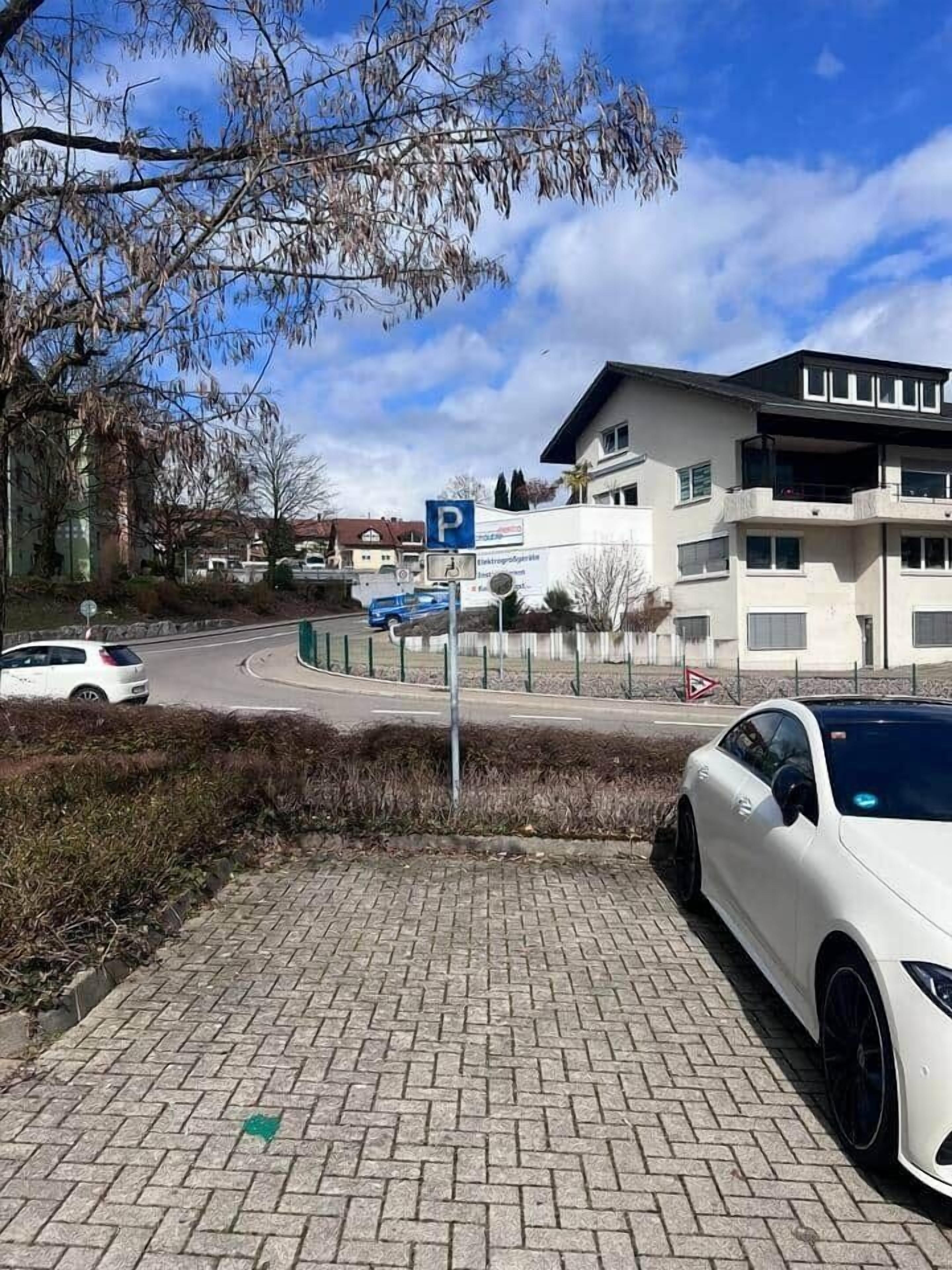 Un área de estacionamiento con un coche blanco aparcado a la derecha, un letrero de estacionamiento azul y un edificio con varias ventanas y balcones al fondo.