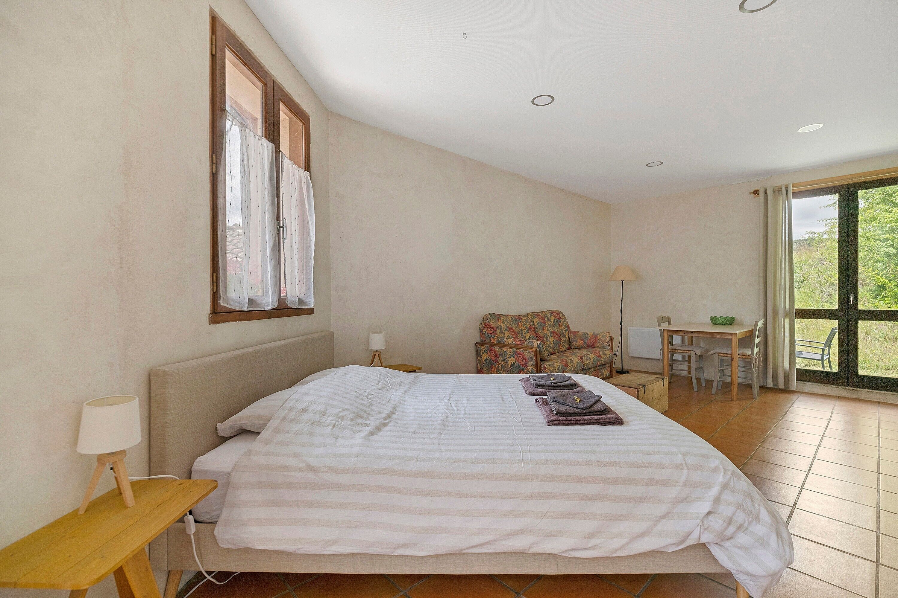 Un dormitorio con cama, una mesita, una silla, un ventanal y vistas al exterior.