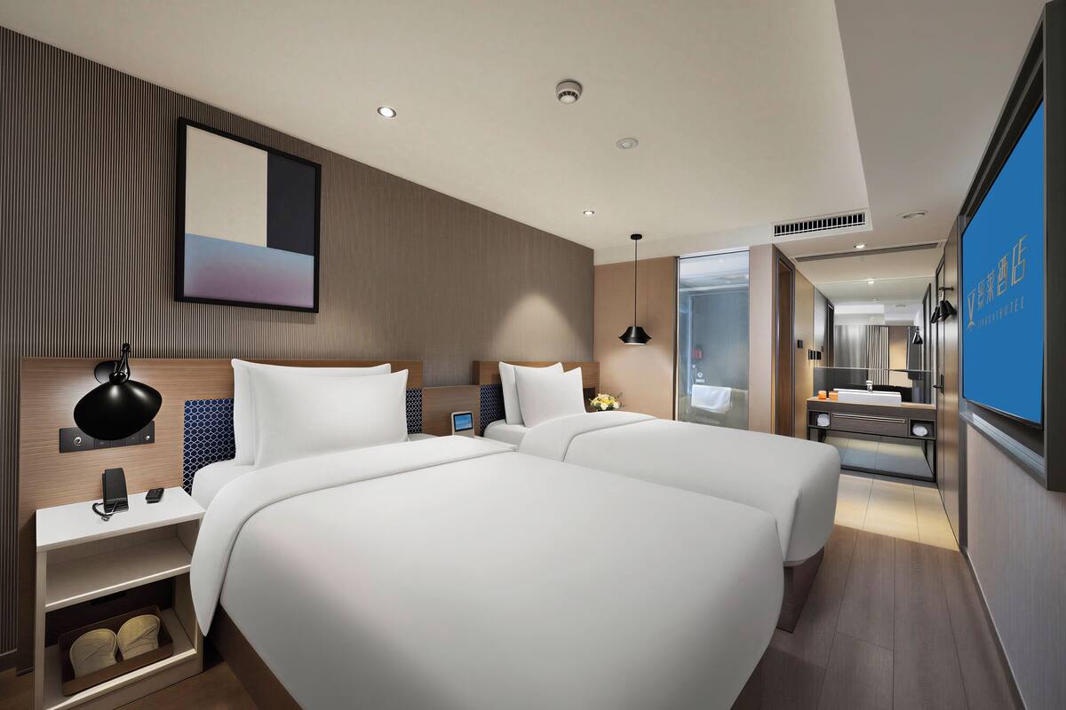 A modern hotel room with two beds, a bedside table with a lamp, a small bedside table with a phone, and a large mirror.