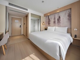 A hotel room with a large bed, a desk, a chair, a wardrobe, a shower, and a wall-mounted light fixture.