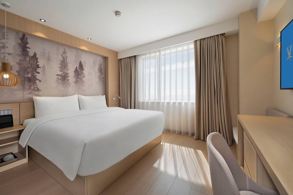 A modern hotel room with a large bed, a desk, a chair, a desk lamp, and a large window with curtains.