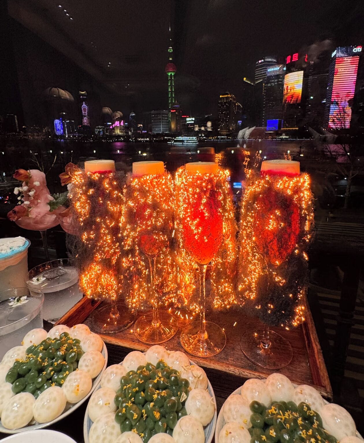 Een tafel met drankjes en snacks, met op de achtergrond een stadshemelbeeld en een toren met verlichte lampen.