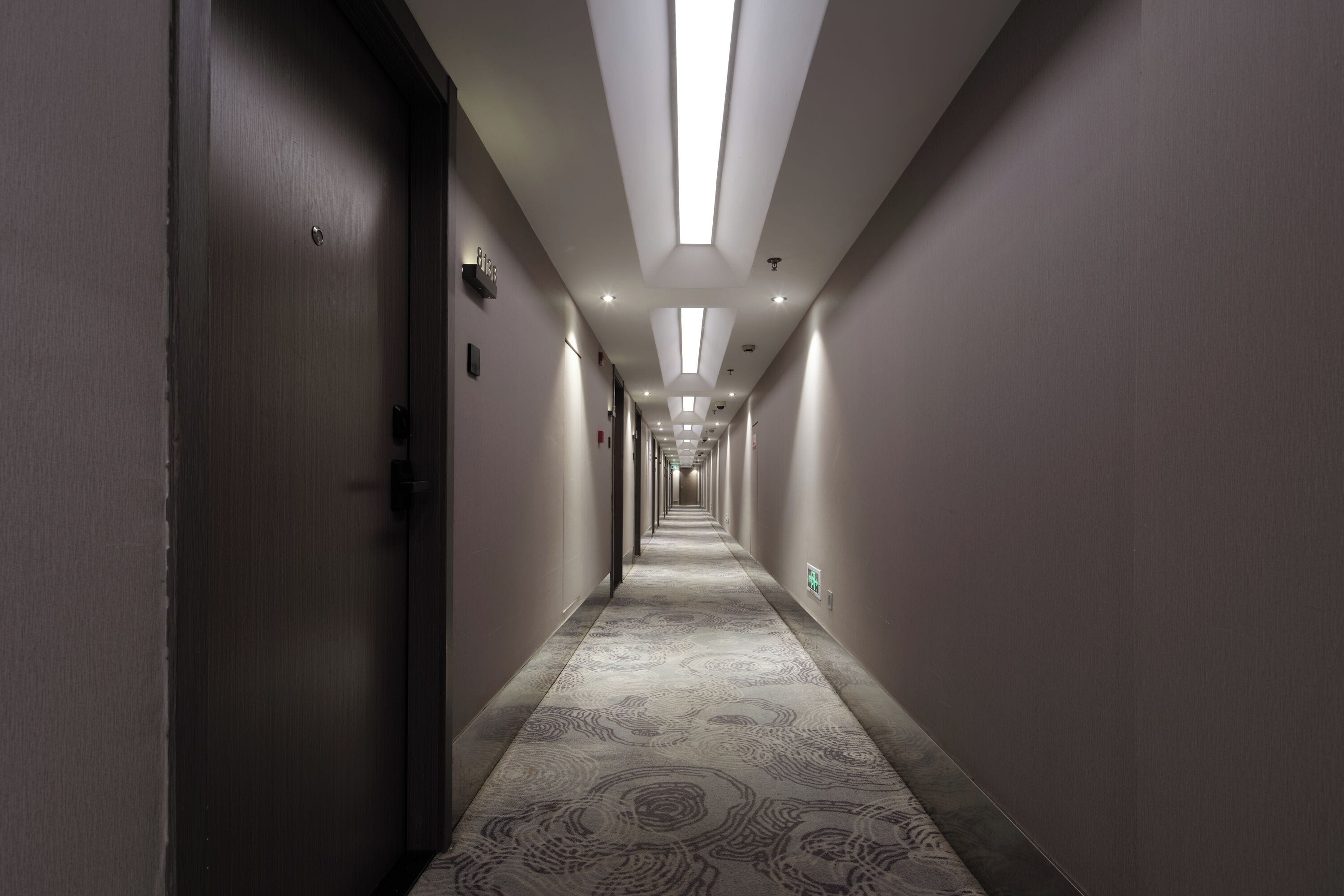 A long, narrow hotel corridor with a patterned carpet, recessed lighting, and doors on both sides.