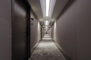 A long, narrow hotel corridor with a patterned carpet, recessed lighting, and doors on both sides.