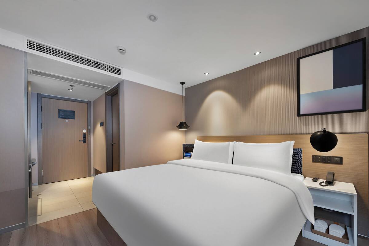 A modern hotel room with a large bed, a bedside table with a lamp, a built-in shelf, and a wall-mounted control panel.