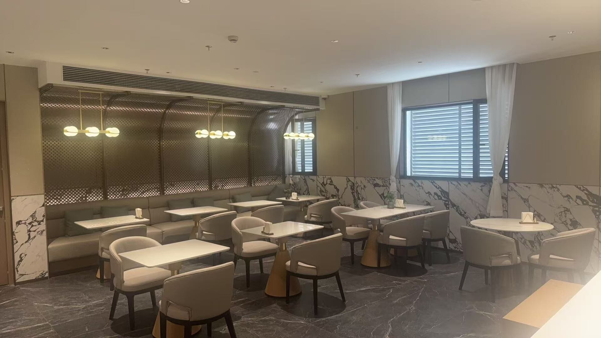 A modern restaurant interior with marble flooring, multiple dining tables and chairs, a seating area with a cushioned bench, and a large window with blinds.