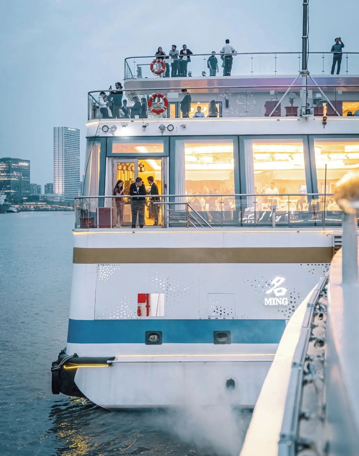 Um ferry com área de convés, boia salva-vidas e o horizonte urbano ao fundo.