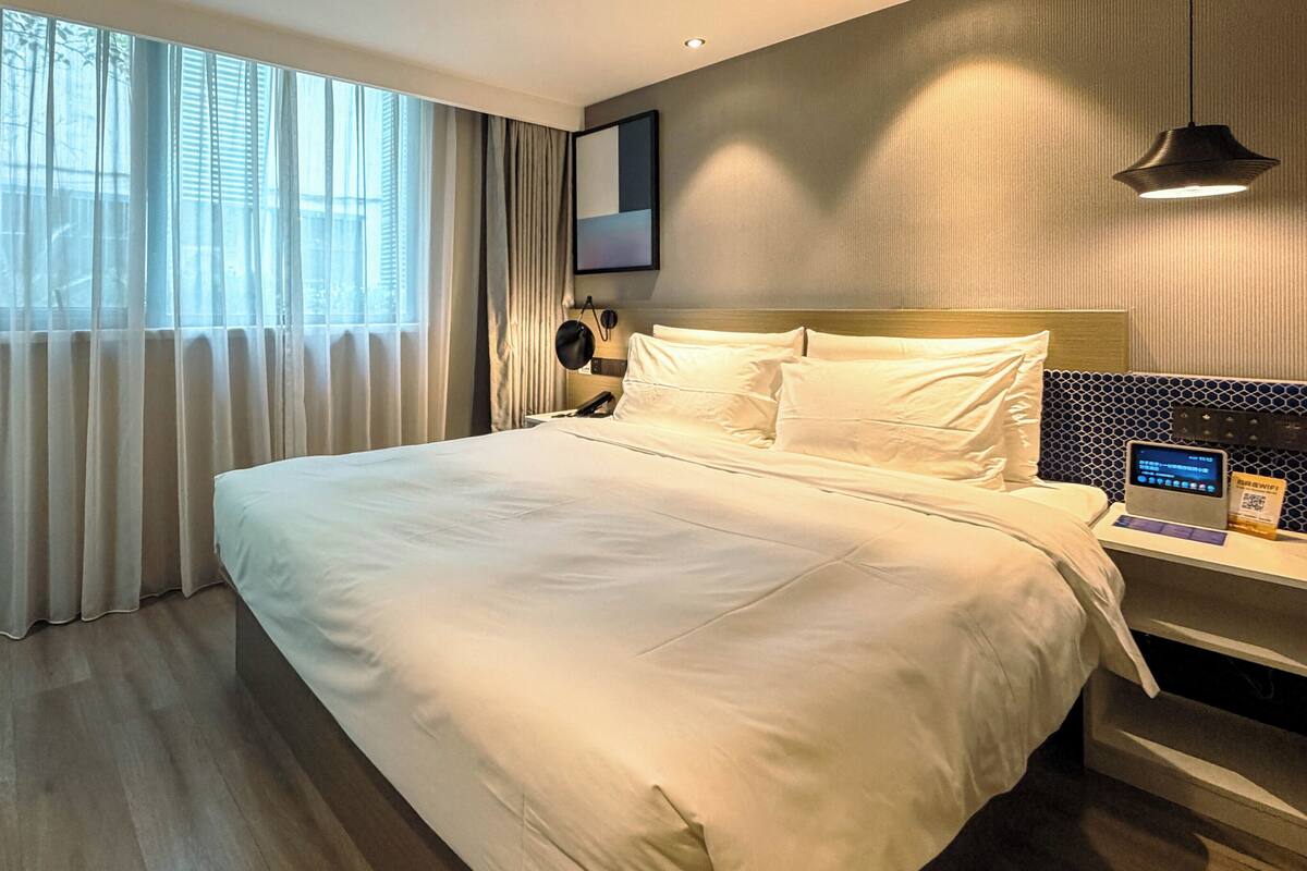 A hotel room with a large bed, a bedside table with a phone and a digital device, a window with curtains, and a wall-mounted light fixture.
