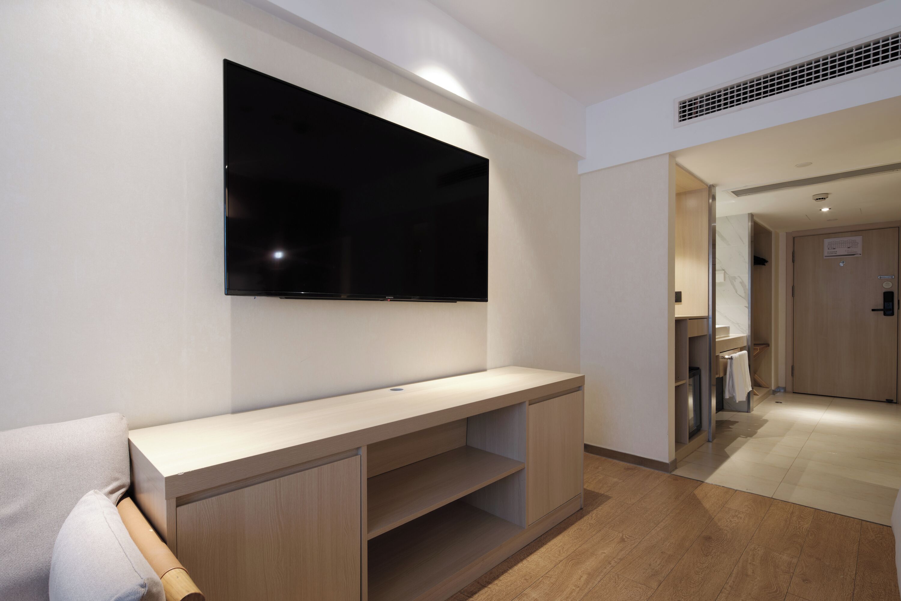 A modern hotel room with a flat-screen TV mounted on the wall, a wooden console table, and a visible bathroom with a towel rack.