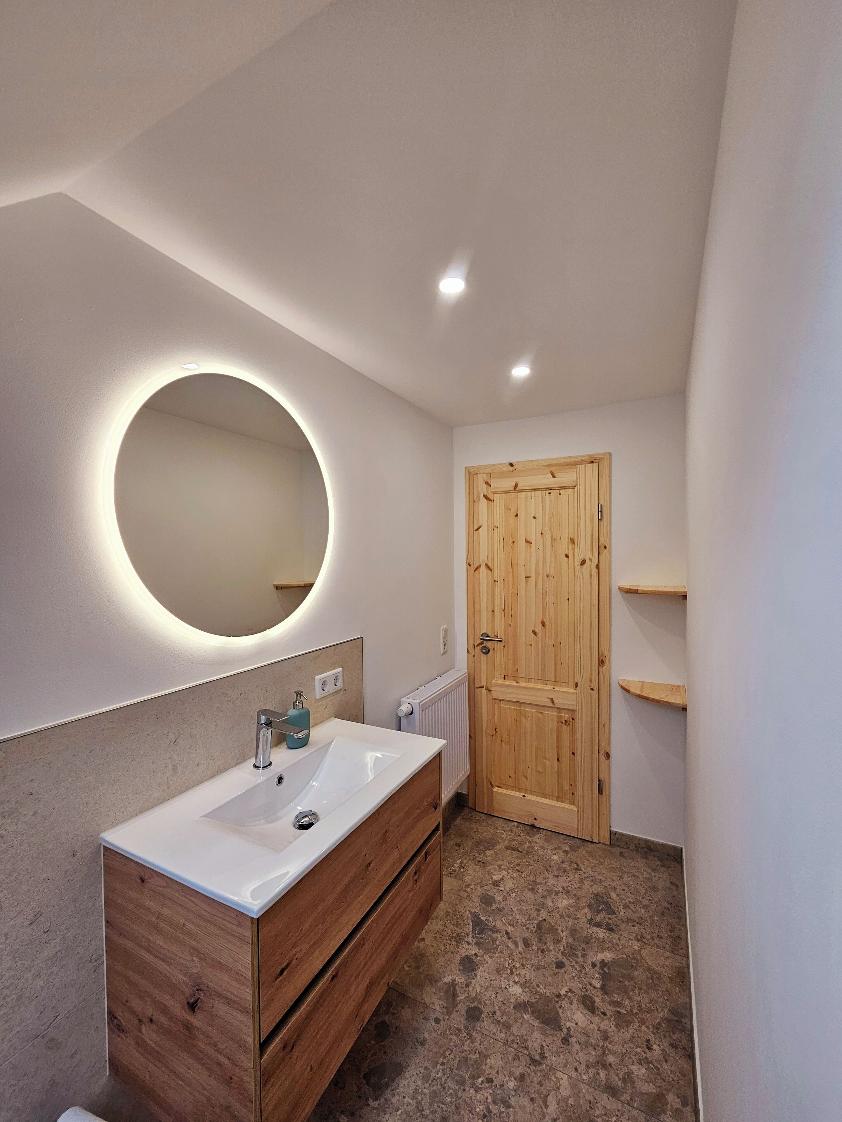 A bathroom with a wooden vanity, a round mirror with integrated lighting, a white sink, and a wooden door.