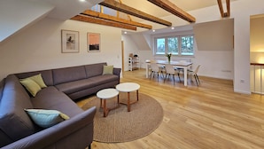 A modern loft with exposed wooden beams, a brown leather sofa, a dining area with white chairs, and a view through a large window.