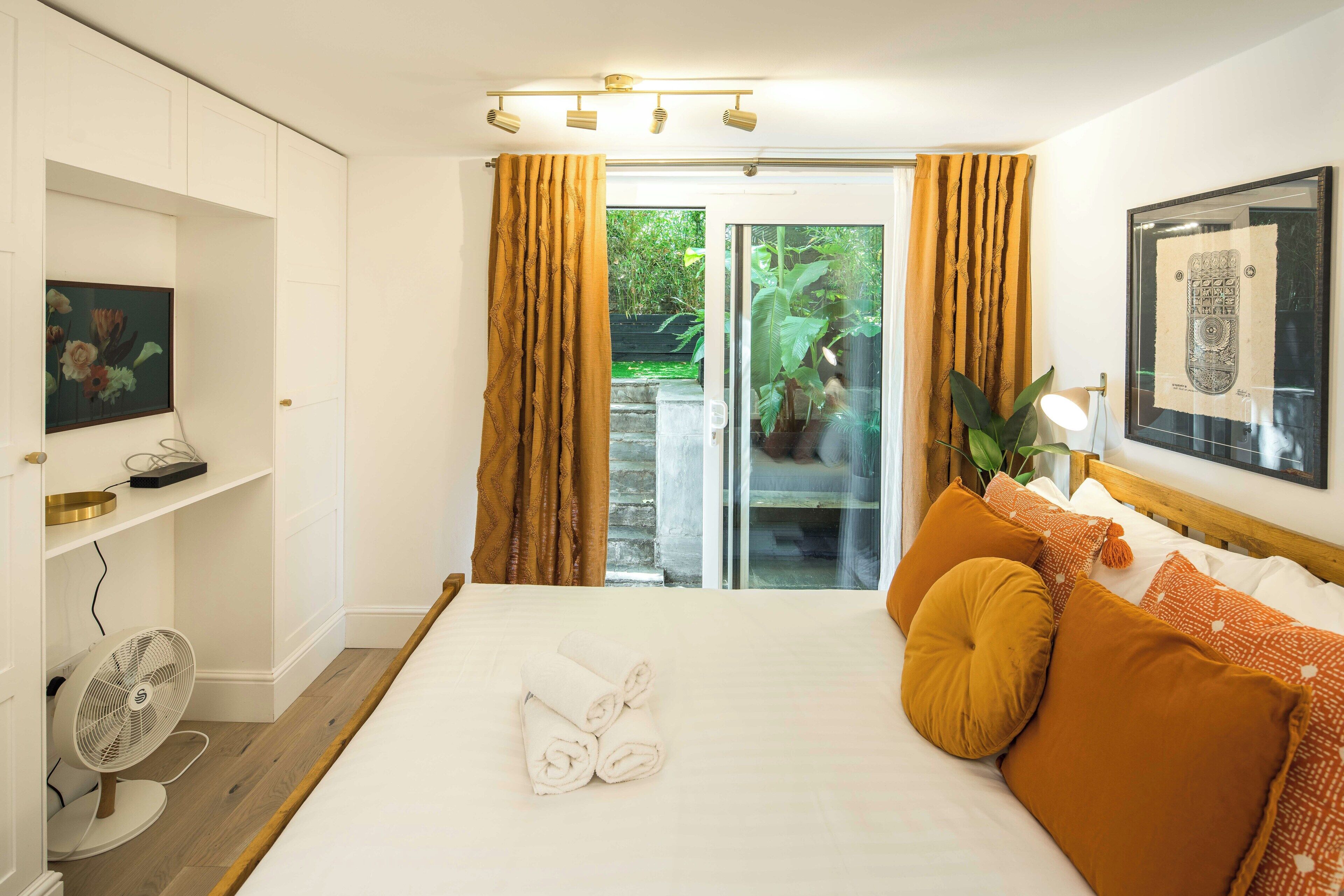 A hotel room with a bed, a TV, a fan, and a view of greenery outside.
