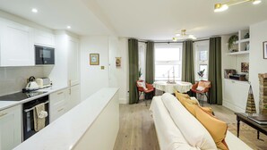 Een moderne keuken met witte kasten, een inbouwoven en een magnetron. Er is een eethoek met een ronde tafel en stoelen, en een bank met kussens.