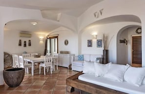 A spacious living area with a white sofa, a wooden coffee table, and a dining table with chairs. There is a large potted plant and a decorative wall piece. - Villa with private pool Porto Cervo (Porto Cervo)