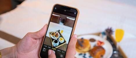 Una persona sostiene un teléfono inteligente que muestra una foto de una bandeja de desayuno con diversos pasteles y bebidas sobre una cama.