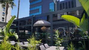 An outdoor seating area with tables and chairs, surrounded by tropical plants, in front of a modern building with large windows.