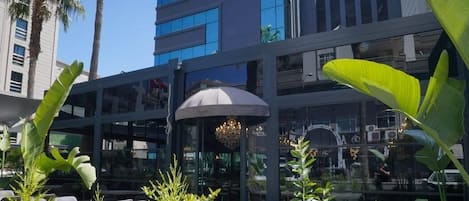 Un espacio al aire libre con mesas y sillas, rodeado de plantas tropicales, frente a un edificio moderno con amplias ventanas.