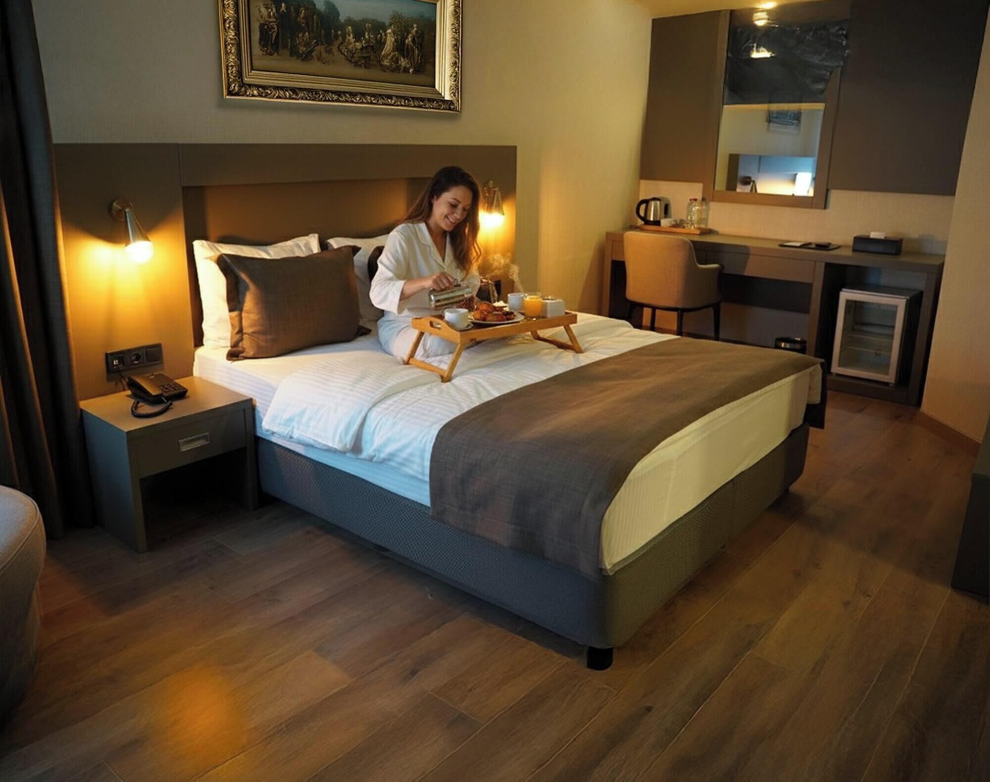 A hotel room with a large bed, a bedside table with a phone, a tray with breakfast items, a painting on the wall, and a desk with a mirror and a lamp.