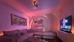 Uma sala de estar moderna com um sofá seccional, uma mesa de centro de vidro, um banquinho de couro branco, uma TV de tela plana e um luminário fixado na parede.